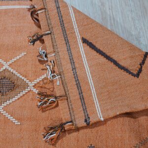 Rust-colored woven rug featuring white and dark geometric patterns, multicolored tassels, resting on wood grain floor.