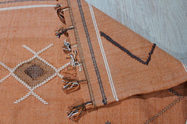 Rust-colored woven rug featuring white and dark geometric patterns, multicolored tassels, resting on wood grain floor.