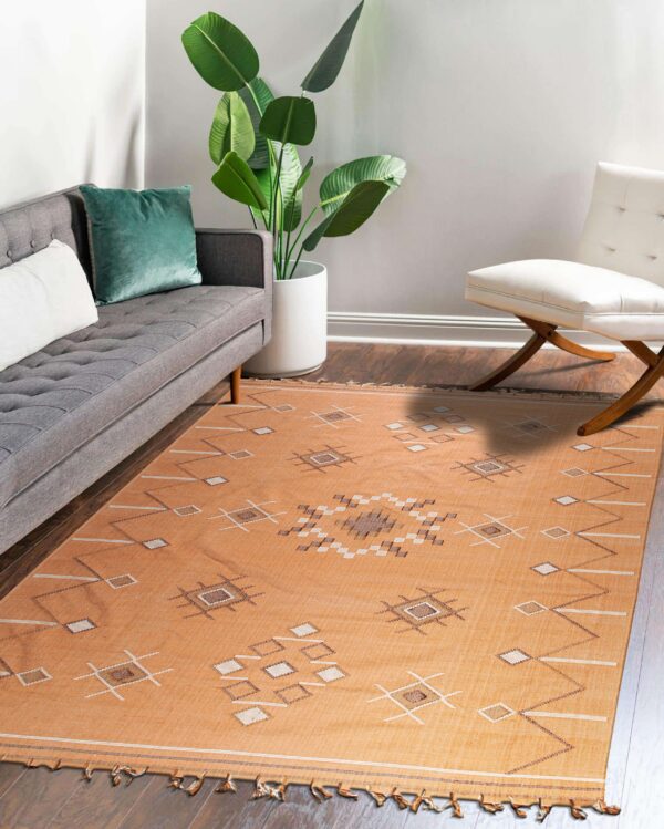 Tan flat-weave rug with geometric patterns on a wood floor between a gray couch and white chair.