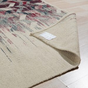 A cream rug with red and teal geometric patterns is folded over, showing the woven backing on light wood flooring.