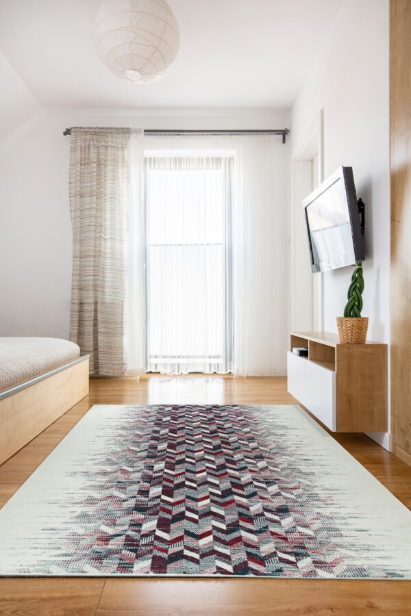 A light cream rug with a central burgundy, blue, and gray geometric chevron pattern lies on a wood floor in a sunny room.