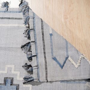 Gray-blue geometric rug with black, white, and blue patterns and tassels folded on light wood flooring.