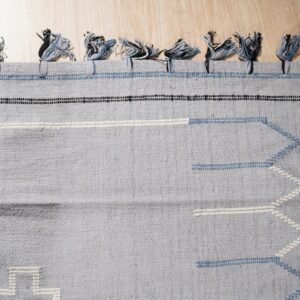Gray-blue flatweave rug with tassels, black and white stripes, and pale blue geometric stitching on wood floor.