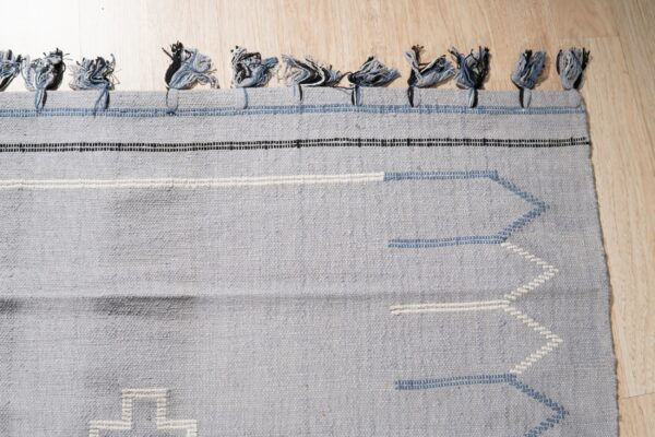 Gray-blue flatweave rug with tassels, black and white stripes, and pale blue geometric stitching on wood floor.