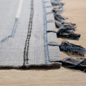 Low pile gray rug displays blue zigzag stitches, vertical stripes, and multi-tone tassels on light wood.