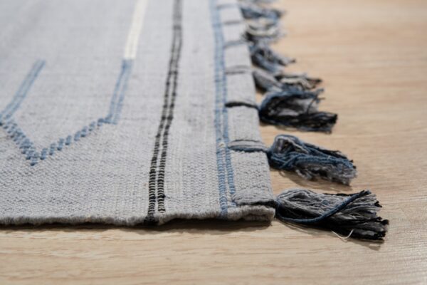 Low pile gray rug displays blue zigzag stitches, vertical stripes, and multi-tone tassels on light wood.