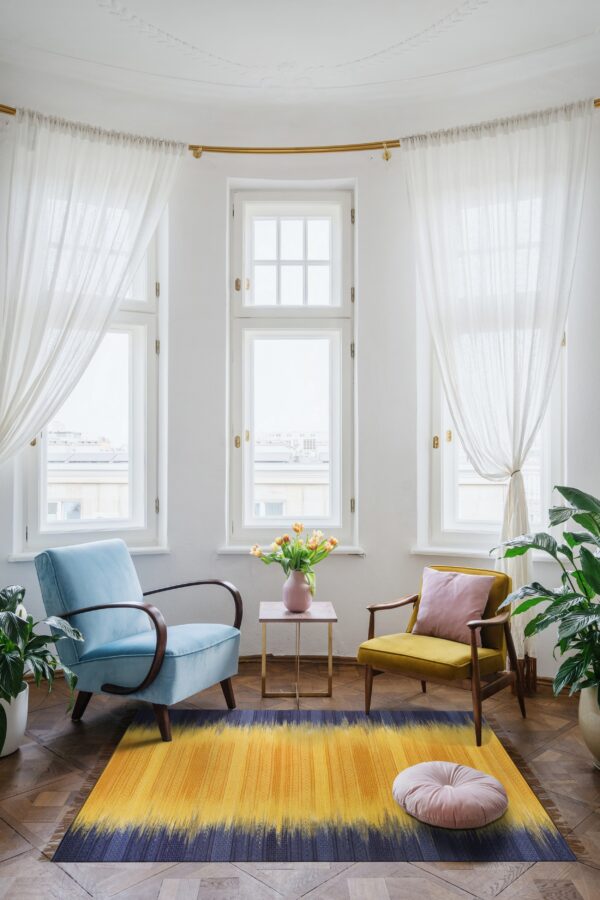 A yellow and blue ikat-style rug anchors a bay window seating area with two vintage velvet chairs on wood floors.