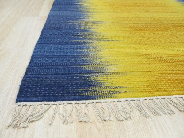 Flatweave rug with a blue to yellow color gradient, subtle patterns, and light fringe on wood flooring.