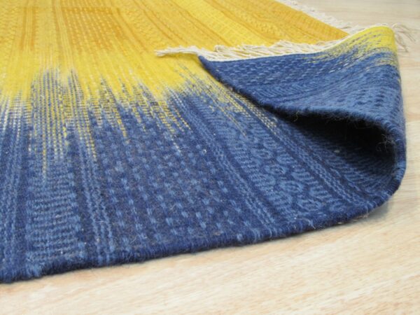 A yellow and indigo rug featuring a vertical fading stripe pattern is partially folded on light wood flooring.