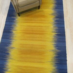 Yellow and blue flatweave rug with abstract fading colors and woven horizontal patterns under a beige upholstered armchair.