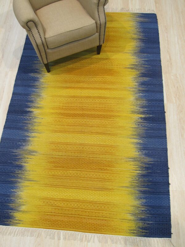 Yellow and blue flatweave rug with abstract fading colors and woven horizontal patterns under a beige upholstered armchair.