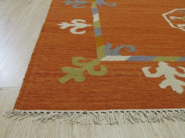Burnt orange flatweave rug corner with white fringe and multicolored stylized border on light wood flooring.