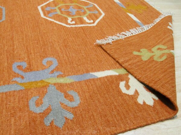 Terracotta low-pile rug with light blue, white, and yellow geometric motifs is folded on light wood flooring.