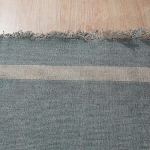 Tightly woven blue rug with a wide beige stripe and fringe, resting on light wood plank flooring.