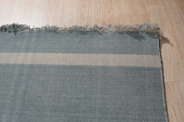 Tightly woven blue rug with a wide beige stripe and fringe, resting on light wood plank flooring.