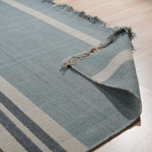 Blue flatweave rug with gray stripes and fringe curled up on light wood plank flooring.