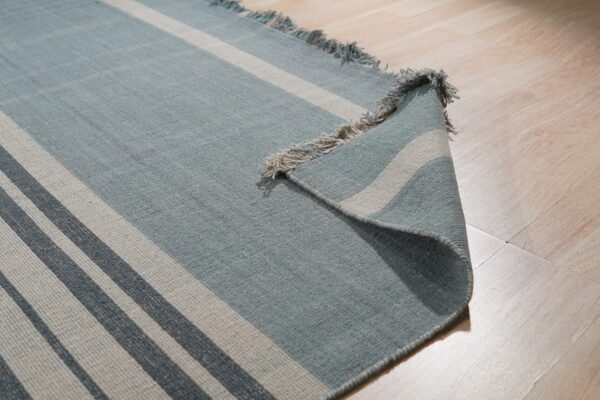 Blue flatweave rug with gray stripes and fringe curled up on light wood plank flooring.