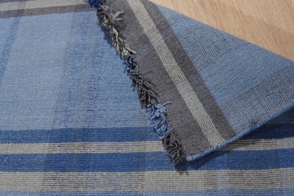 Blue and gray striped flatweave rug is folded over a wooden floor, showing short fringe detail.