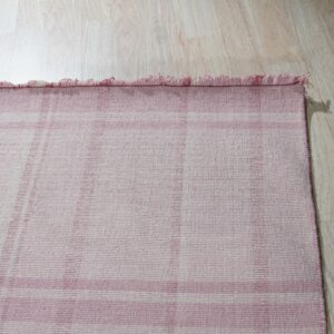 Close-up of a pink and white plaid flatweave rug with short fringes on light wood plank flooring.