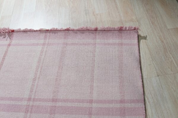 Close-up of a pink and white plaid flatweave rug with short fringes on light wood plank flooring.