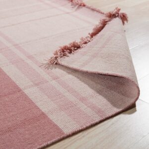 Pink and cream striped flat-weave rug with tassel fringe, partly rolled on light wood plank flooring.
