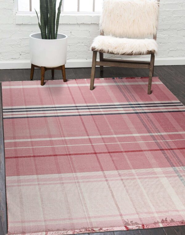A pink tartan rug rests on dark wood flooring next to a fuzzy white chair and a tall snake plant.
