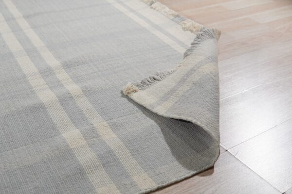 Light gray and cream striped low-pile rug with fringe, partially rolled on light plank flooring.