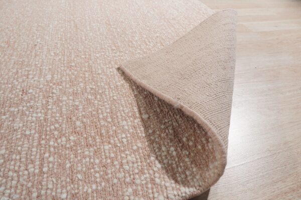 Textured pale pink and white speckled rug partially rolled back, showing the backing on light wood flooring.