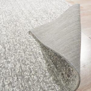 A textured gray and white rug is peeled back to show its woven backing on light wood floor.