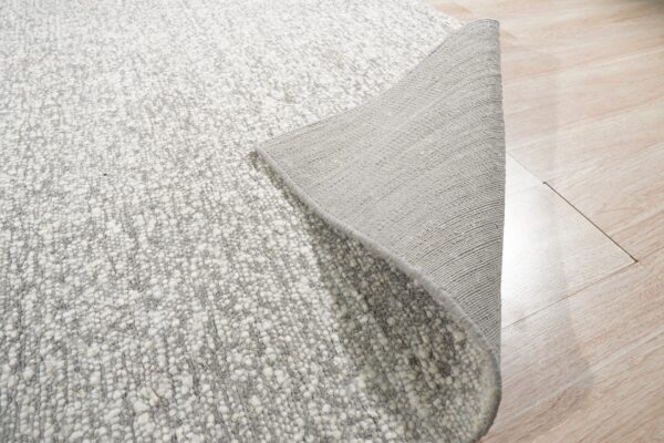 A textured gray and white rug is peeled back to show its woven backing on light wood floor.