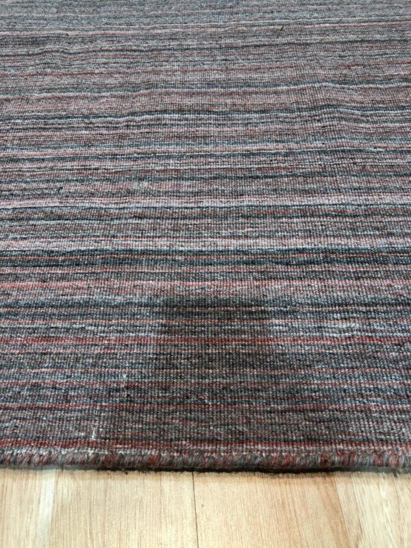 Textured low-pile rug with horizontal bands of charcoal gray, rust, and light blue on light wood flooring.