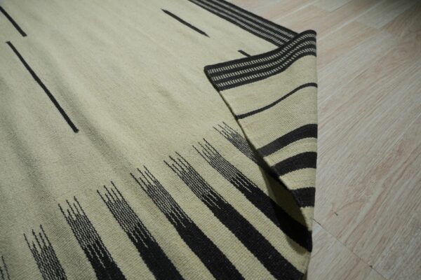 Light-colored flatweave rug with abstract dark stripes and bands displayed on blonde wood flooring.