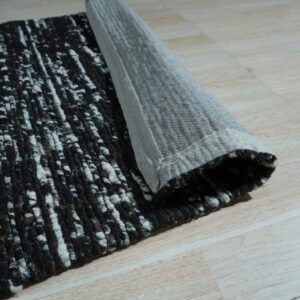 A black and white textured rug is rolled up, revealing its gray woven backing on light wood flooring.
