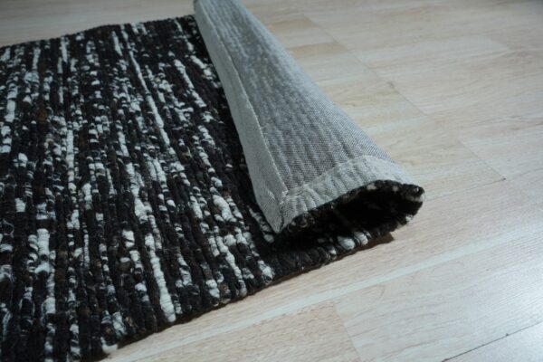 A black and white textured rug is rolled up, revealing its gray woven backing on light wood flooring.