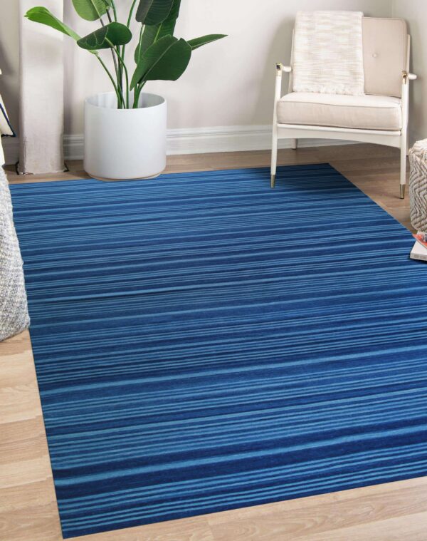 A blue and teal striped rug lies on wood floors next to a white armchair and a potted plant.