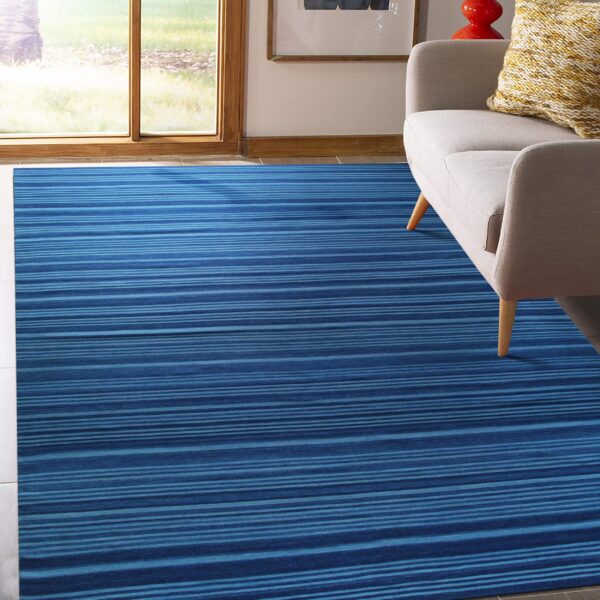 A blue low-pile rug with horizontal stripes sits under a gray sofa near a large sunlit window.