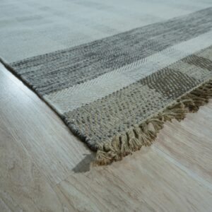 Low-angle view of a striped flatweave rug in cream and natural tones with fringe on light wood.