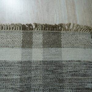 Woven rug detail showing tan fringe and block patterns in beige and gray on light wood planks.