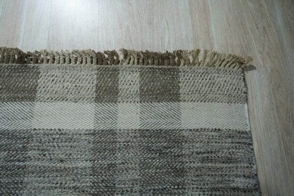 Woven rug detail showing tan fringe and block patterns in beige and gray on light wood planks.