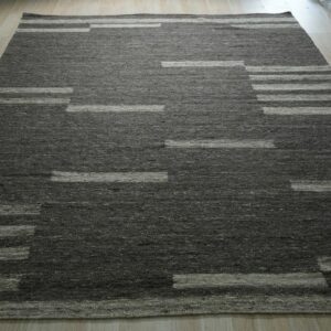 Dark gray geometric rug with light stripes on wood flooring, surrounded by rolled textiles.