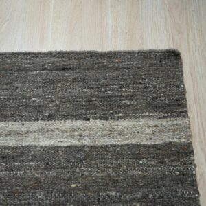 Flatweave rug corner with thick gray and light beige horizontal stripes on light wood planks.