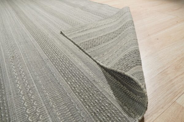 Close-up of a gray flatweave rug featuring textured stripes, partially rolled on light blonde wood flooring.