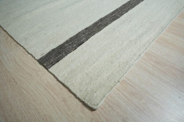 Close view of a nubby, cream-colored rug featuring a thick charcoal stripe on pale wood planks.