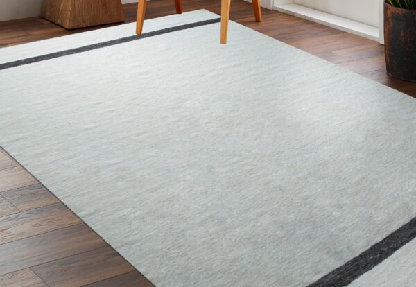 Light gray low-profile rug with black lines on dark wood floors near a wooden chair leg.