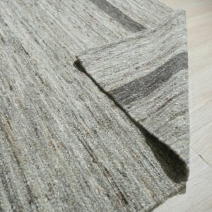Close-up of a folded, chunky, heathered gray rug with dark stripes lying on light wood flooring.