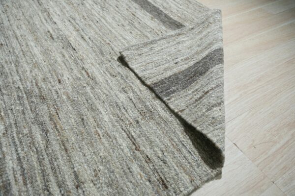 Close-up of a folded, chunky, heathered gray rug with dark stripes lying on light wood flooring.