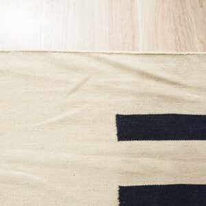 Low-pile beige rug with dark geometric stripes rests on light wood grain flooring.