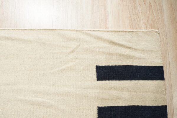 Low-pile beige rug with dark geometric stripes rests on light wood grain flooring.