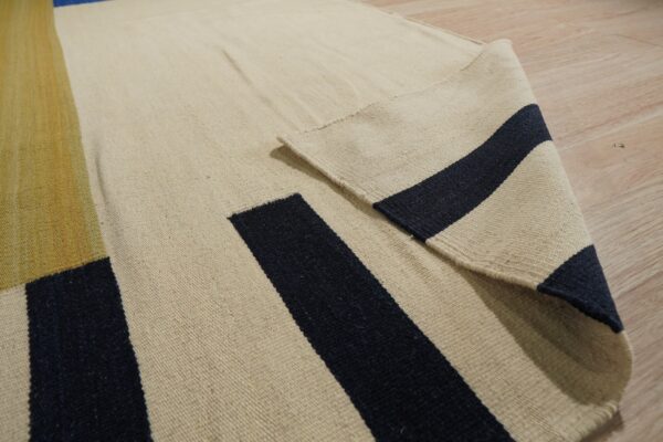 Flatweave rug in beige, navy, and gold geometric stripes folded on light wood floor.