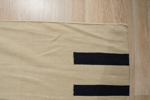 Low-profile beige rug with a black geometric block pattern lying on light blonde wood flooring.
